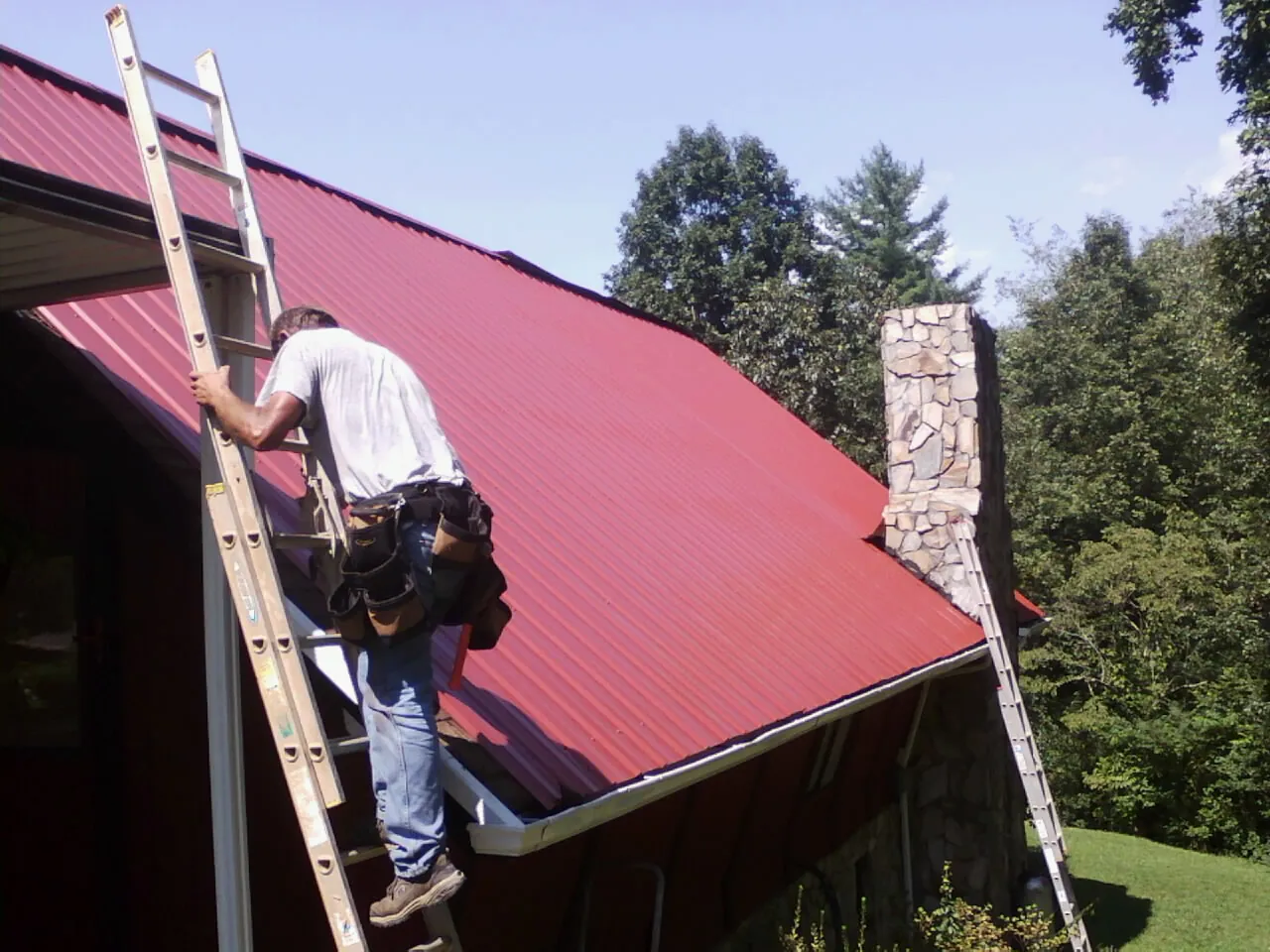 Roofer on a ladder beside a red metal roof and stone chimney — Soffit & Fascia Repair by Roofing Brothers in Kittery Point